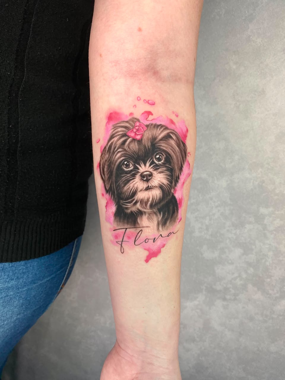 Tatuagem realista de shih tzu com aquarela rosa e o nome Flora no antebraço
