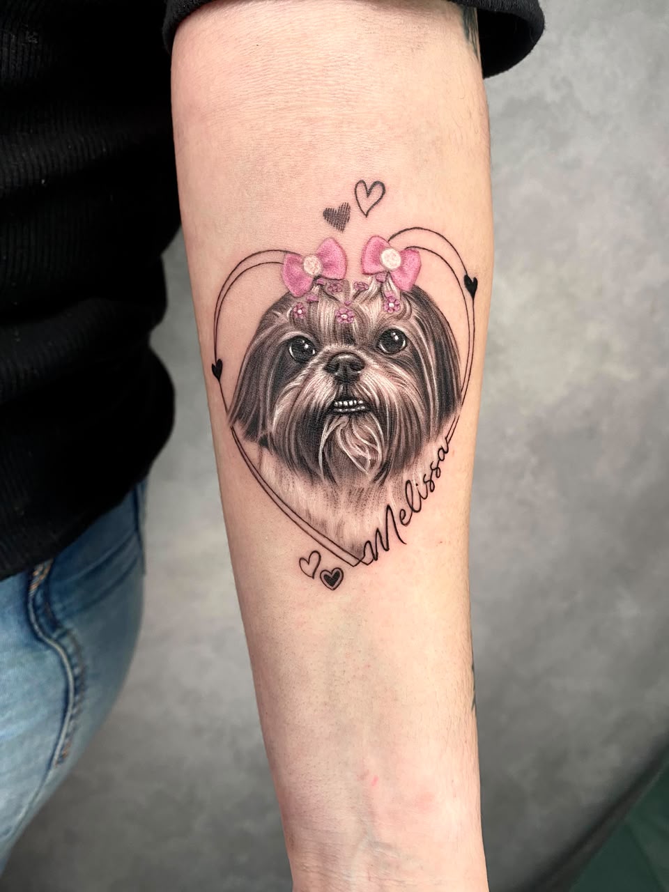 Tatuagem realista de shih tzu com laços rosa, coração e o nome Melissa no antebraço